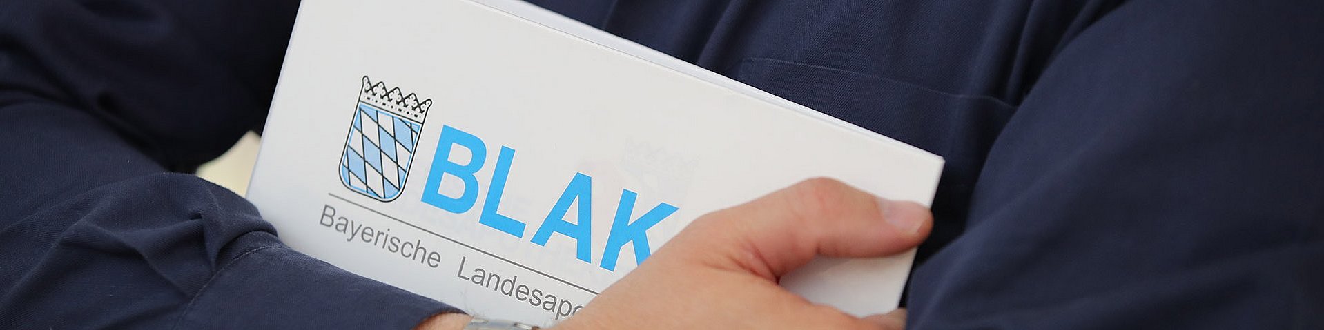 Jemand mit blauem Hemd und einer silbernen Armbanduhr am Arm hält einen Ordner der BLAK in den Händen. Auf dem Ordner ist das BLAK-Logo sehr groß abgebildet.