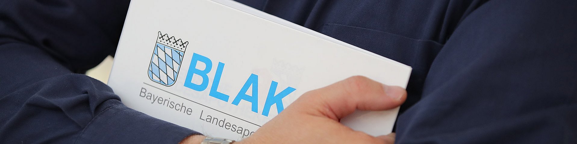Ordner der Bayerischen Landesapothekerkammer Jemand mit blauem Hemd und einer silbernen Armbanduhr am Arm hält einen Ordner der BLAK in den Händen. Auf dem Ordner ist das BLAK-Logo sehr groß abgebildet.