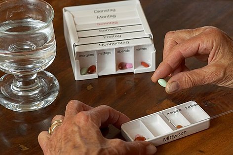 Eine Patientin hält eine Tablette zwischen ihren Fingern. Vor ihr liegen die Medikamentenfächer der einzelnen Wochentage und ein Glas Wasser steht auf dem Tisch.