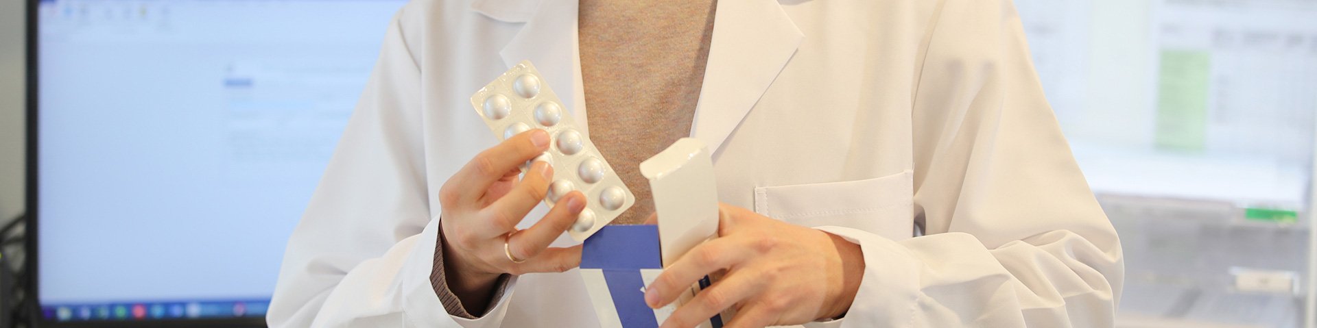 Ein Apotheker mit weißem Kittel entpackt eine Medikamentenschachtel und holt die verplisterten Tabletten heraus. Im Hintergrund sieht man einen Bildschirm.