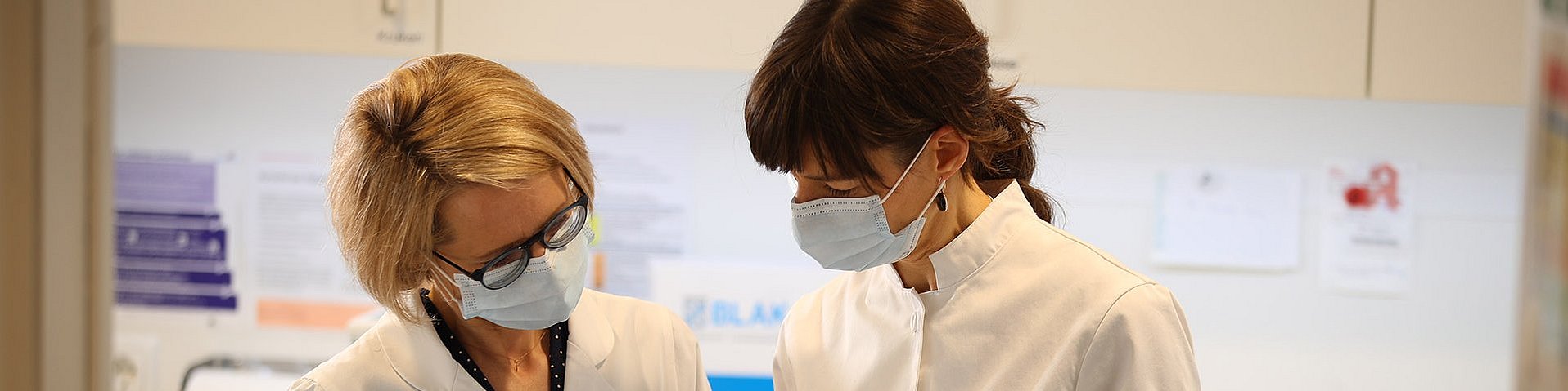Zwei Apothekerinnen mit Mundschutz sehen sich konzentriert Formulare an. Sie befinden sich in einem Labor. Eine hält eine weiß-rote Dose in den Händen.