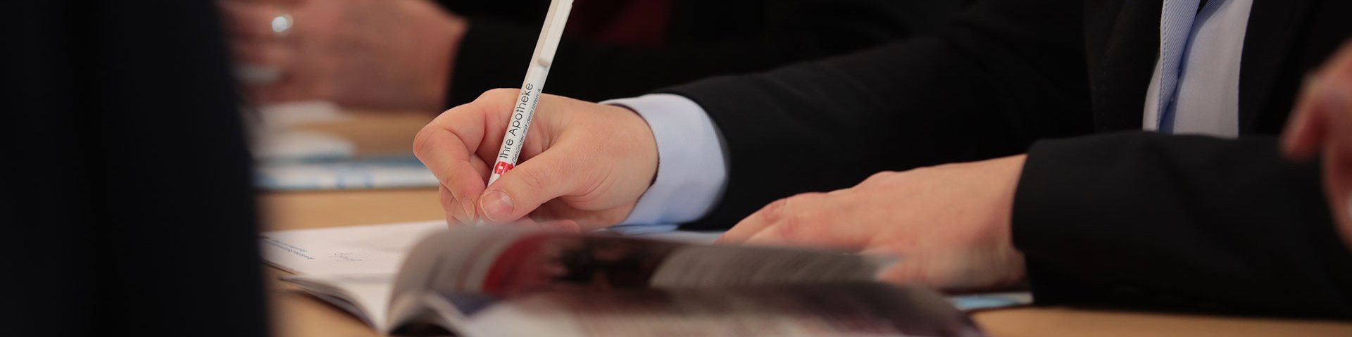 Meeting in der Bayerischen Landesapothekerkammer. Mitarbeiter sitzen mit ihren Unterlagen am Besprechungstisch. Eine Person macht sich Notizen in ihr Heft.