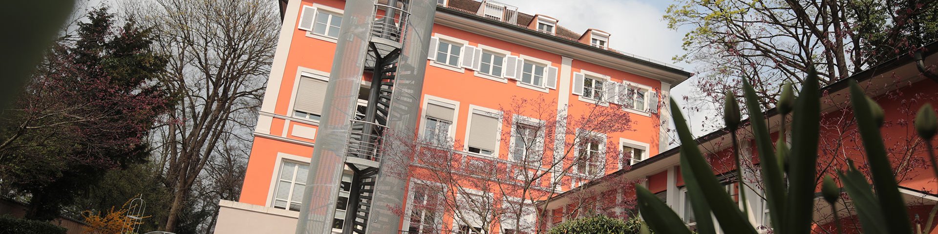 Das Gebäude der Bayerischen Landesapothekerkammer in der Außenansicht. Es ist orange mit weiß umrahmten Fenstern und einer Notfallwendeltreppe am Haus. 