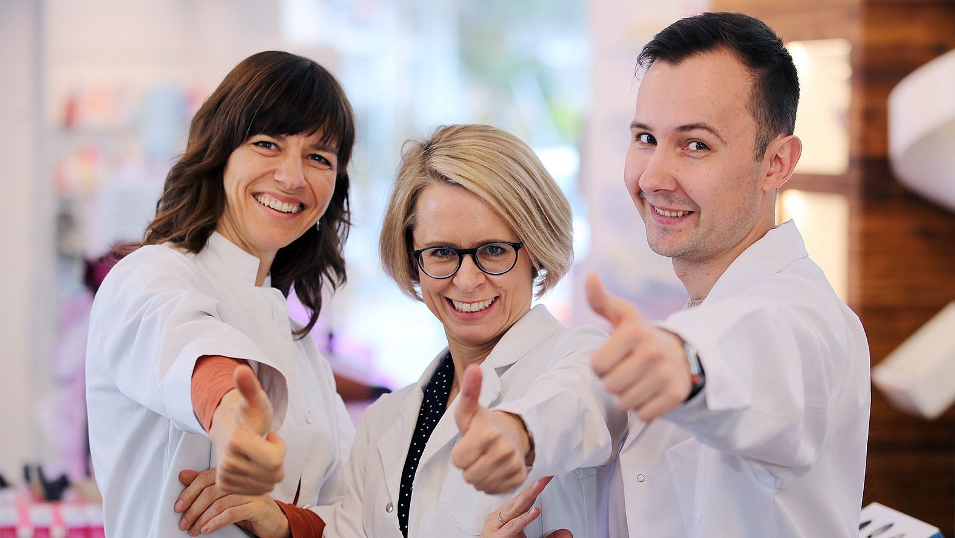 Gruppenbild von BLAK Apotheker Drei Apotheker der BLAK stehen als Gruppe in der Apotheke. Alle drei lachen und zeigen mit dem Daumen nach oben. Hinter ihnen stehen die Medikamentenregale.
