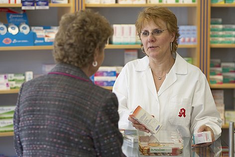 Eine Apothekerin berät eine Kundin. Dabei stehen sie vor den Medikamentenregalen.